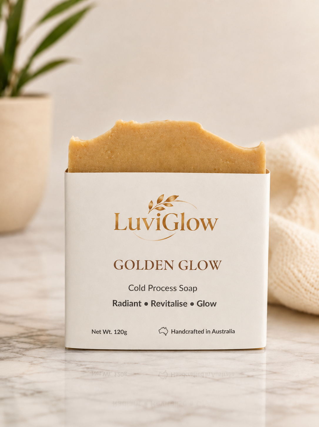 A bar of LuviGlow Golden Glow cold process soap is shown partially unwrapped from its white paper packaging, set on a marble surface with a soft beige background and a blurred plant.