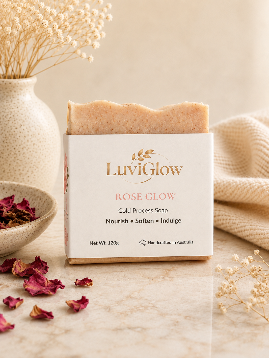 A bar of LuviGlow Rose Glow cold process soap in its white and gold packaging, styled on a marble surface with dried flowers, a ceramic vase, and scattered rose petals.